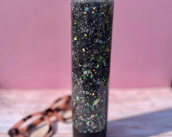 May include: A clear glass cylinder filled with black glitter and iridescent green and silver stars. The cylinder has a black lid and is sitting on a light wood surface.