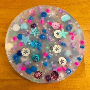 May include: A clear round resin coaster with a variety of colorful and glittery embellishments, including seashells, starfish, and beads.