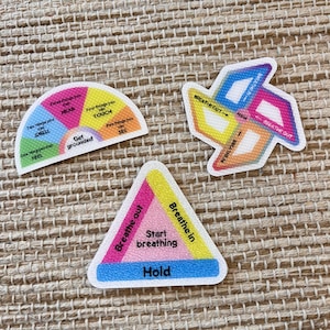 May include: Three colorful stickers with grounding and breathing exercises. One semi-circle labeled Get grounded, one pinwheel shape with BREATHE IN and BREATHE OUT text, and one triangle labeled Start breathing with Hold and Breathe out text.