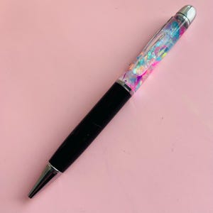 Könnte beinhalten: Ein schwarzer und silberner Kugelschreiber mit einem transparenten Schaft, der mit bunten Glitzer und Konfetti gefüllt ist. Der Stift hat eine silberne Spitze und einen silbernen Aufsatz. Der Stift liegt auf einer rosa Oberfläche.
