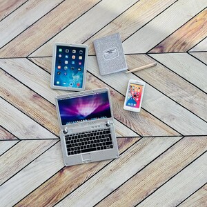 May include: A silver laptop computer with a black keyboard and a white screen displaying the Apple logo and the words "Mini Book". A silver tablet computer with a black screen displaying a grid of colorful icons. A silver smartphone with a black screen displaying a grid of colorful icons. A gray notebook with the text "Composition Book" printed on the front. A wooden surface with a chevron pattern.