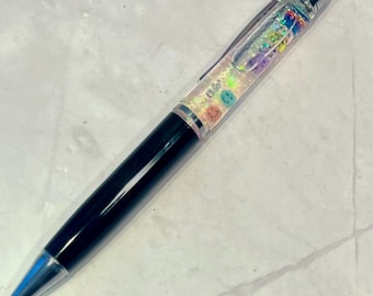 May include: A black pen with a clear barrel filled with colorful glitter and smiley faces. The pen has a silver clip and a silver cap.