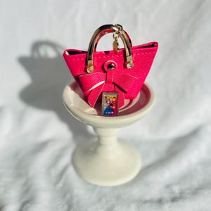 May include: A pink faux leather handbag keychain with a gold handle and a small phone charm. The keychain is on a white pedestal.
