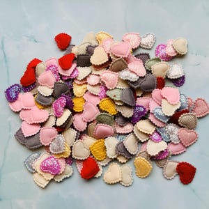 以下が含まれることがあります： 赤、ピンク、グレー、黄色、ベージュなど、さまざまな色のハート型の布製装飾品のコレクション。キラキラしたアクセントが付いているハートもあります。ハートは水色の表面に散らばっています。