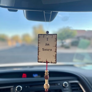 May include: A wooden car charm with the words "I Am Yours" printed on it. The charm is rectangular with a distressed border and hangs from a red string with wooden beads. The charm is hanging from a car's rearview mirror.