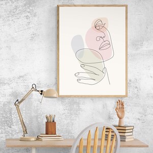 May include: A framed line art print of a face with abstract shapes in pink, green, and purple. The print is hanging on a white wall above a desk with a lamp, a chair, and a stack of books.