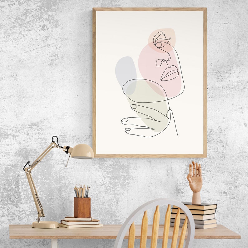 May include: A framed line art print of a face with abstract shapes in pink, green, and purple. The print is hanging on a white wall above a desk with a lamp, a chair, and a stack of books.