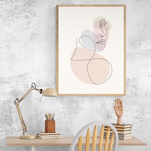 May include: A framed line art print of a face with pink and purple abstract shapes. The print is hanging on a white wall above a desk with a lamp, a wooden pencil holder, books, and a wooden chair.