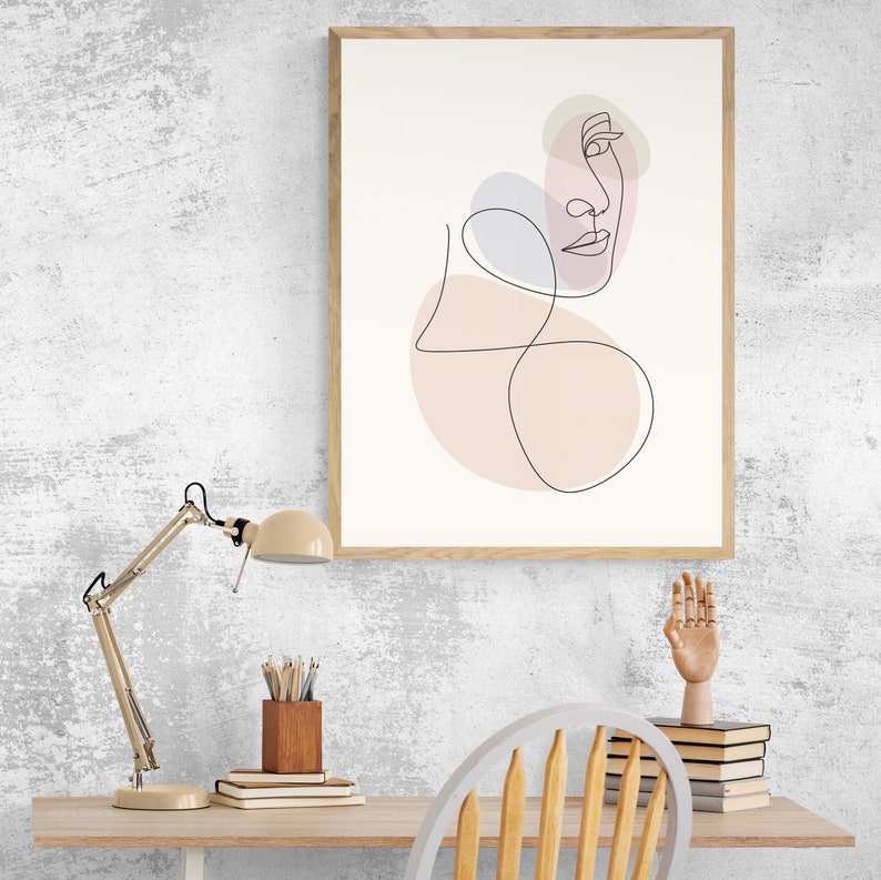 May include: A framed line art print of a face with pink and purple abstract shapes. The print is hanging on a white wall above a desk with a lamp, a wooden pencil holder, books, and a wooden chair.