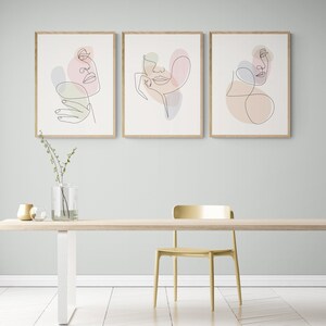 May include: Three framed line art prints of abstract faces with pastel colored shapes. The prints are hung on a light blue wall above a wooden table with a gold vase and a gold bowl.