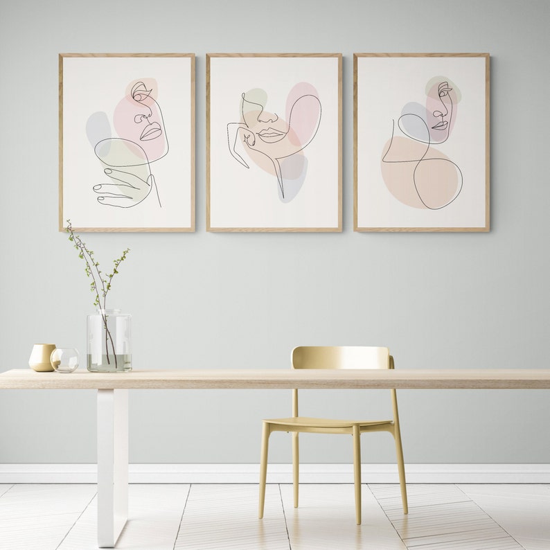 May include: Three framed line art prints of abstract faces with pastel colored shapes. The prints are hung on a light blue wall above a wooden table with a gold vase and a gold bowl.