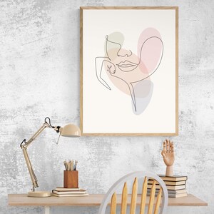 May include: A framed line drawing of a face with pastel colored shapes behind it. The drawing is on a white background and the frame is light wood. The drawing is hanging on a white wall.