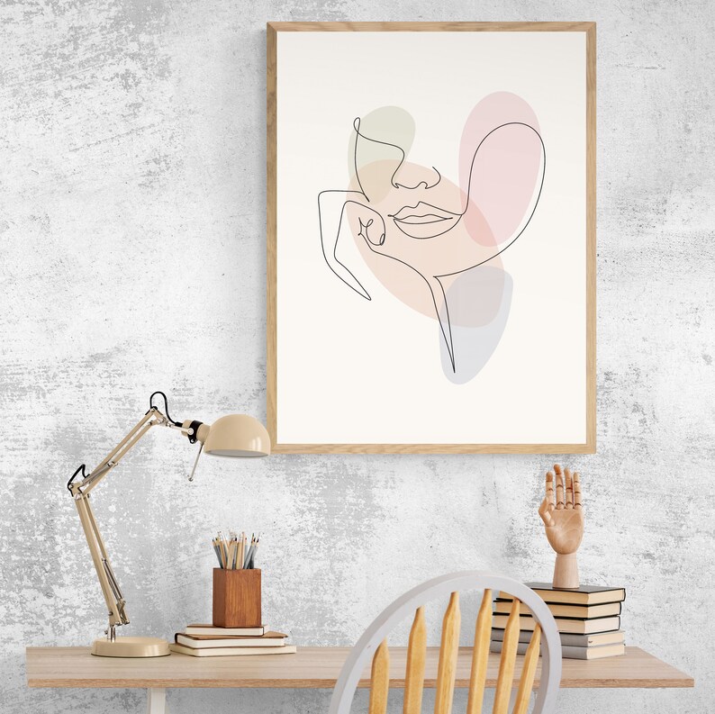 May include: A framed line drawing of a face with pastel colored shapes behind it. The drawing is on a white background and the frame is light wood. The drawing is hanging on a white wall.