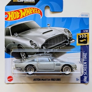 Könnte beinhalten: Ein silbernes Hot Wheels Druckgussmodell eines Aston Martin 1963 DB5, verpackt auf einer blau-weißen Karte. Die Karte trägt den Text "Goldfinger Sixty Years" und "HW Screen Time". Das Auto ist im Maßstab 1:64.