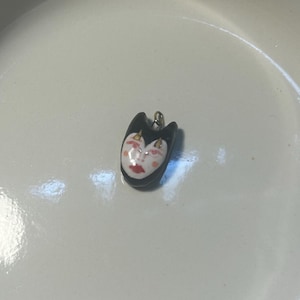 May include: A small pendant shaped like a stylized face with a black outer border, a white center, and painted red lips and pink cheeks. The pendant features a small metal loop at the top for attachment, resting inside a white bowl with a black rim.