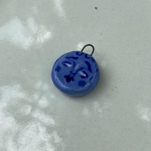 May include: A small, round, blue pendant with a smiling face design. The pendant has a silver-colored loop at the top for attaching to a chain or cord. The pendant is sitting on a white ceramic surface.