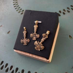 Può includere: Tre pendenti a forma di croce color bronzo esposti su un espositore nero. Le croci presentano intricati dettagli floreali e figurativi. L'espositore è posto su una superficie azzurra a motivi.