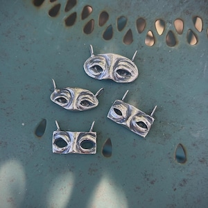 May include: Four silver-toned, eye-shaped pendants with small, raised details. Each pendant has two small, upward-pointing protrusions. The pendants are arranged on a teal-colored, textured surface with teardrop-shaped cutouts.