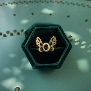 May include: A gold-colored ring with a butterfly wing design, set in a dark green velvet hexagonal ring box. The ring has an openwork design and a central stone. The background is a light green surface.