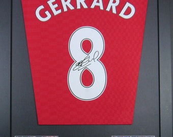 Könnte beinhalten: Gerahmtes rotes Fußballtrikot mit dem Namen "GERARD" und der Nummer "8" in Weiß. Das Trikot ist von Steven Gerrard signiert. Der Rahmen enthält zwei Fotos von Gerrard beim Fußballspielen.