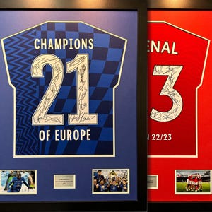 Pode incluir: Duas camisas emolduradas, uma azul com o número 21 e o texto "Champions of Europe", e outra vermelha com o número 3 e o texto "Arsenal 22/23". Ambas as camisas têm assinaturas.