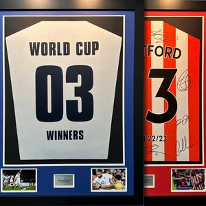 Op de afbeelding: Twee ingelijste sportshirts. Eén is wit met de tekst "WORLD CUP 03 WINNERS". De andere is rood en wit gestreept met nummer 3. Beide in zwarte lijsten met respectievelijk blauwe en rode passe-partouts. Inclusief kleine foto's.