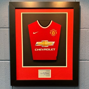 Pode incluir: Uma camisa de futebol vermelha enquadrada com o logotipo do Manchester United e o logotipo da Chevrolet. A camisa é exibida em um fundo preto com um tapete vermelho. O texto "My First Manchester United" está em um rótulo branco abaixo da camisa.