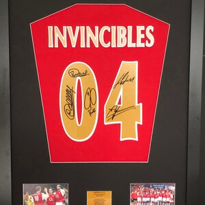 Op de afbeelding: Een ingelijst rood Arsenal voetbal shirt met het woord "INVINCIBLES" en het nummer "04" in goud. Het shirt is gesigneerd door verschillende spelers. De lijst bevat ook twee kleinere foto's van het team.