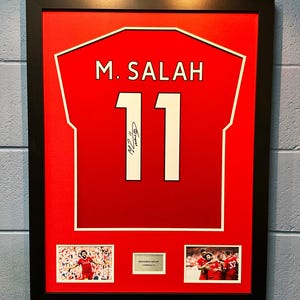 Pode incluir: Uma camisa de futebol vermelha enquadrada com o nome "M. SALAH" e o número "11" em branco. A camisa é assinada com tinta vermelha. Há duas fotos menores abaixo da camisa, uma de um jogador comemorando um gol e a outra de um grupo de jogadores comemorando.