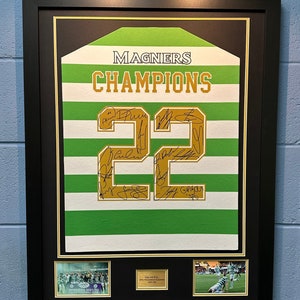 May include: A framed green and white jersey with the number 22 in gold and the words "MAGNERS CHAMPIONS" in gold. The jersey is signed by multiple players. There are two smaller photos below the jersey, one of a group of people in green and white jerseys and the other of a football match.