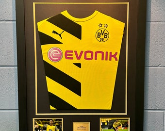 May include: A framed yellow and black football shirt with black diagonal stripes. The shirt has the Puma logo, the text "EVONIK" and the BVB 09 logo. The frame includes two smaller photos of a footballer in action.