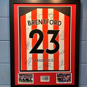 Könnte beinhalten: Ein gerahmtes rot-weiß gestreiftes Trikot mit der Nummer 23 und dem Wort "BRENTFORD" in Schwarz. Das Trikot ist von mehreren Spielern signiert und enthält den Text "SEASON 22/23". Der Rahmen enthält zwei kleinere Fotos des Teams.