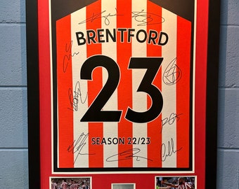 May include: A framed red and white striped jersey with the number 23 and the word "BRENTFORD" in black. The jersey is signed by multiple players and includes the text "SEASON 22/23". The frame includes two smaller photos of the team.
