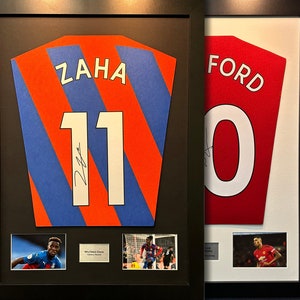 May include: Two framed soccer jerseys, one blue and orange with the name "ZAHA" and number "11" and the other red with the name "FORD" and number "10". Both jerseys have a player's signature and a small photo of the player in action.