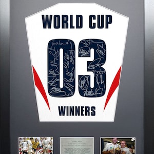 Pode incluir: Uma camisa de rugby branca emoldurada com o número "03" e as palavras "WORLD CUP WINNERS" nela. A camisa é assinada pela equipe vencedora da Copa do Mundo de Rugby da Inglaterra de 2003. O quadro também inclui duas fotos da equipe.