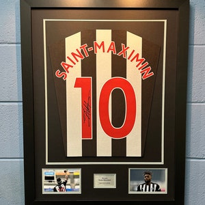 May include: A framed black and white jersey with the name "SAINT-MAXIMIN" and the number "10" in red. The jersey is signed by Allan Saint-Maximin. The frame includes two smaller photos of Saint-Maximin playing football.