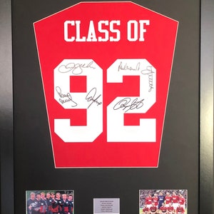 Può includere: Una maglia rossa incorniciata con il numero 92 e il testo "CLASS OF" in bianco. La maglia è firmata da più persone. Ci sono due foto più piccole sotto la maglia, una di un gruppo di persone in uniformi bianche e nere e l'altra di un gruppo di persone in uniformi rosse e bianche.
