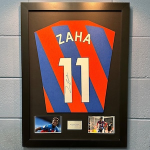 Peut inclure: Un maillot de football encadré et signé avec le nom "ZAHA" et le numéro "11" au dos. Le maillot est rouge et bleu avec des rayures blanches. Deux photos plus petites du joueur sont incluses dans le cadre.