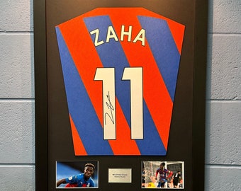 May include: A framed, signed football shirt with the name "ZAHA" and the number "11" on the back. The shirt is red and blue with white stripes. Two smaller photos of the player are included in the frame.
