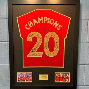 Może przedstawiać: Ramka z czerwonym strojem z napisem "CHAMPIONS" i numerem "20" w złotych literach. Strój jest podpisany przez wielu graczy. W ramce znajdują się dwa mniejsze zdjęcia zespołu świętującego.
