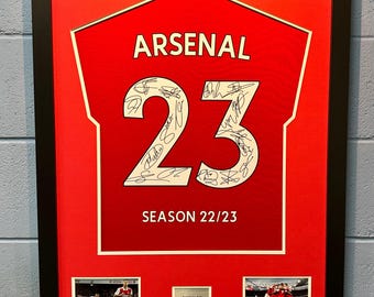 May include: A framed red and white Arsenal football shirt with the number 23 and the text "SEASON 22/23". The shirt is signed by multiple players. There are two smaller photos of Arsenal players below the shirt.