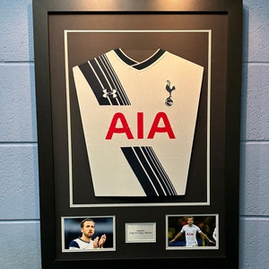 Puede incluir: Una camiseta de fútbol blanca enmarcada con detalles en negro y azul. La camiseta tiene la palabra "AIA" impresa en rojo en la parte delantera. La camiseta está enmarcada con un marco negro e incluye dos fotos más pequeñas de un jugador de fútbol vistiendo la camiseta.