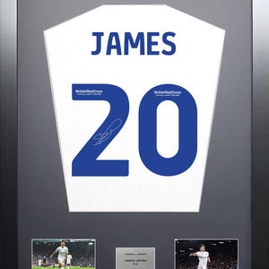 May include: A framed white soccer jersey with the name "JAMES" and the number "20" in blue. The jersey is signed by Daniel James, a player for Leeds United FC. The frame includes two smaller photos of James playing soccer.