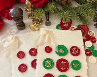 May include: Two off-white handmade paper cards with frayed edges, each tied with a cream ribbon. The cards are decorated with various red and green wax seals featuring gold-embossed holiday motifs like Christmas trees, holly, and candy canes.