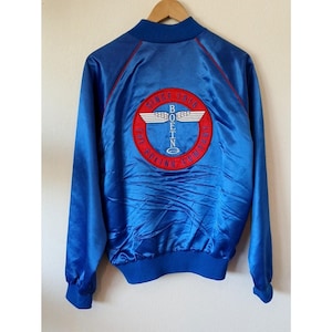May include: A royal blue satin bomber jacket featuring a large circular embroidered patch on the back. The patch displays the text SINCE 1916 THE BOEING COMPANY in red with a white winged logo and the word BOEING in the center. It has red piping.