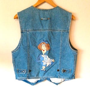 May include: A blue denim vest featuring a sleeveless design with a back yoke and adjustable side tabs. The back panel displays a central embroidered applique of a figure with reddish-brown hair, wearing a blue outfit and holding a patterned quilt.