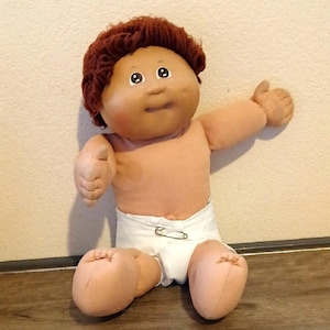 May include: A vintage doll with a soft, tan body, brown curly hair, and wide eyes. The doll is wearing a white diaper secured with a safety pin and is posed with one arm raised in a thumbs-up gesture. The doll is sitting on a wooden surface.
