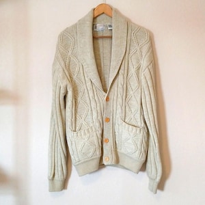 May include: A cream-colored cable knit cardigan featuring a wide shawl collar, long sleeves with ribbed cuffs, and a button-down front closure. The garment includes two front patch pockets and a textured diamond pattern across the front panels.