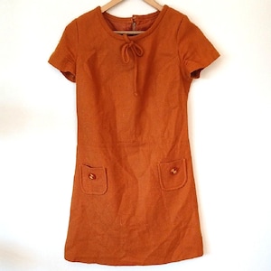 May include: A burnt orange vintage dress with short sleeves, a round neckline, and two front pockets. The dress has a tie detail at the neckline and wooden buttons on the pockets. The dress is hanging on a wooden hanger.