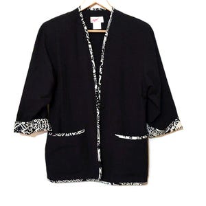 May include: A black open-front jacket featuring a white geometric pattern along the collar, cuffs, and hem. The garment includes two front pockets with matching white patterned trim and a label at the neck with the text Tapa in pink lettering.